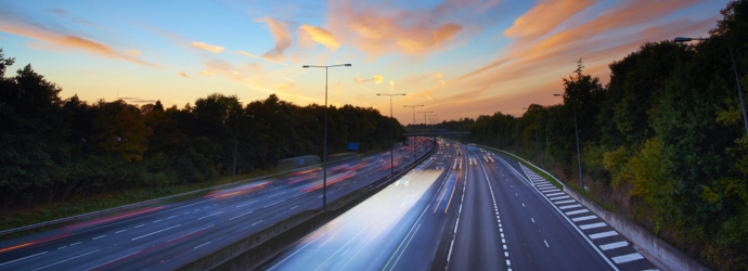 A sunset sky over a busy highway with streaks of car lights. Trees line the road, and the colors evoke a serene yet dynamic atmosphere of evening transit.