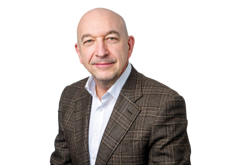 A bald man with a goatee wearing a checked brown suit and white shirt, posed against a white background, slightly smiling.